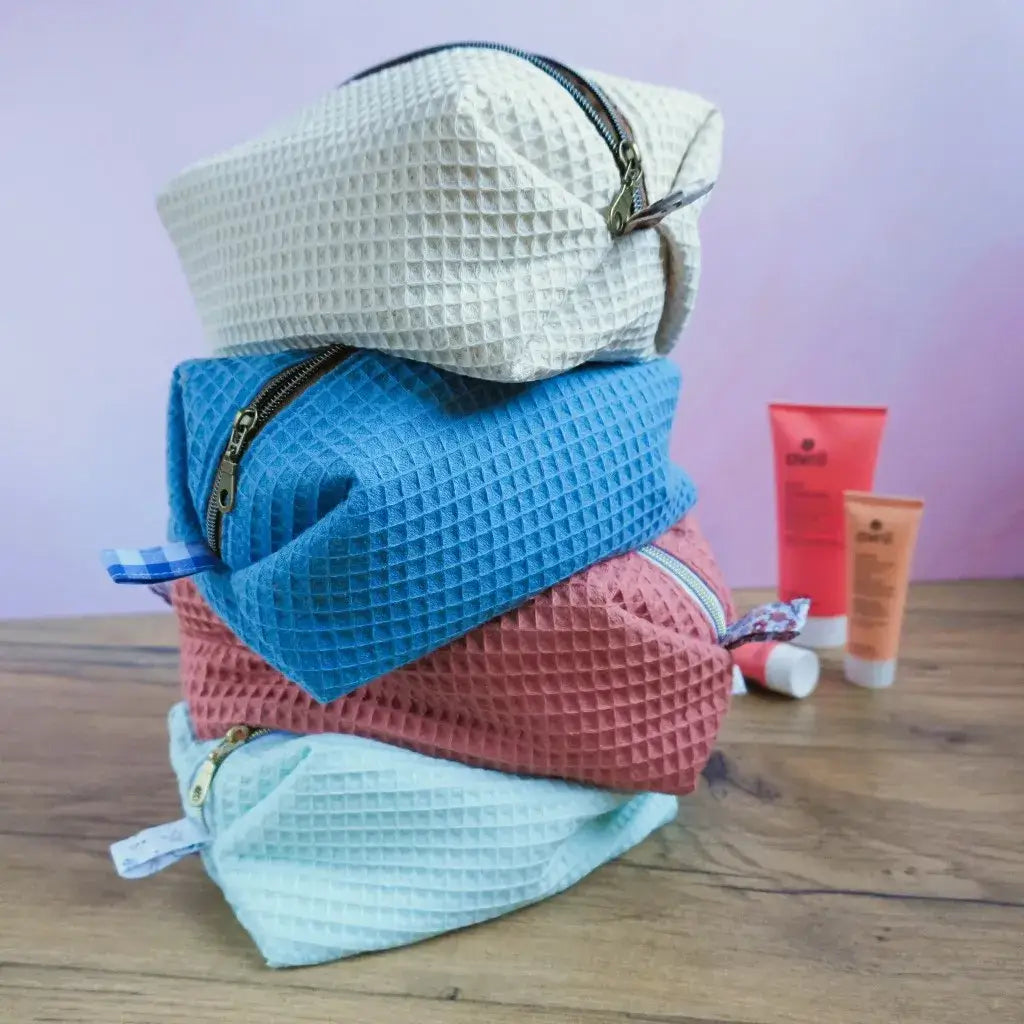 Empilement de quatre trousses carrées en tissu nid d’abeille, déclinées en coloris naturel, bleu denim, marsala et vert menthe. Disposées sur une table en bois avec produits de soin en arrière-plan, elles illustrent une collection artisanale élégante et coordonnée. Chaque trousse présente une fermeture éclair métallique et une doublure imprimée unique. Accessoires faits main parfaits pour le rangement, les voyages ou un coffret cadeau harmonieux.