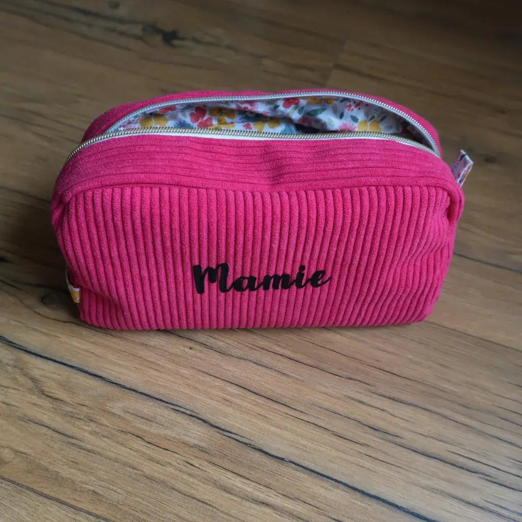 Trousse de toilette en velours côtelé framboise, personnalisée avec l’inscription “Mamie” floquée en velours noir sur le devant. Elle est équipée d’une fermeture éclair argentée et doublée d’un tissu intérieur à motifs floraux colorés. Posée sur un sol en bois, cette trousse artisanale allie praticité et élégance, idéale pour ranger produits de beauté, accessoires ou effets personnels lors des déplacements.