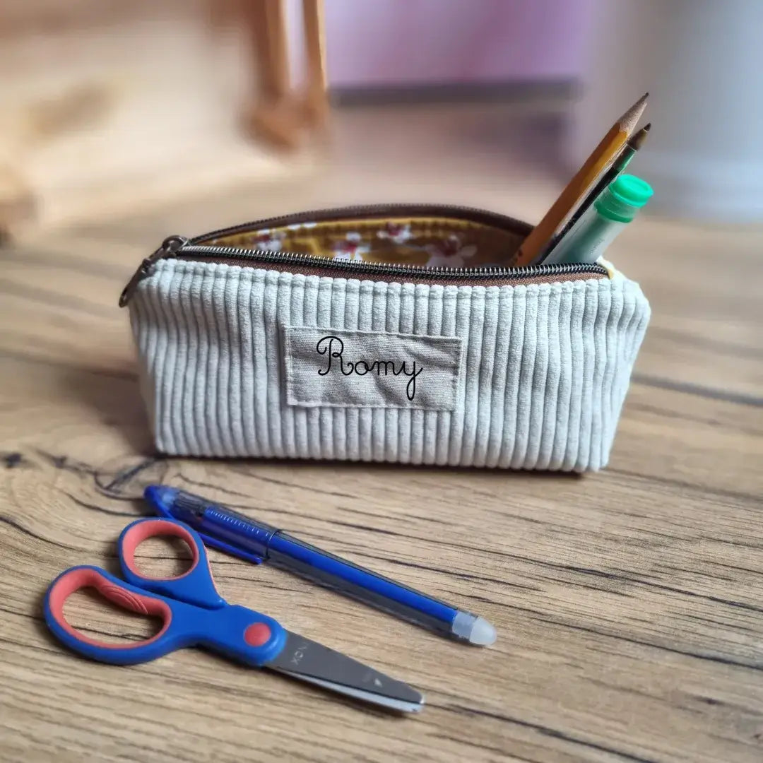 Trousse d’école personnalisée en velours côtelé beige, floquée du prénom "Romy", posée sur un bureau en bois. La trousse est ouverte et contient des crayons et un tube de colle. Devant, une paire de ciseaux pour enfant bleue et rouge ainsi qu’un stylo bille bleu. Intérieur doublé d’un tissu en coton enduit fleuri. Accessoire scolaire pratique et élégant, idéal pour la rentrée.