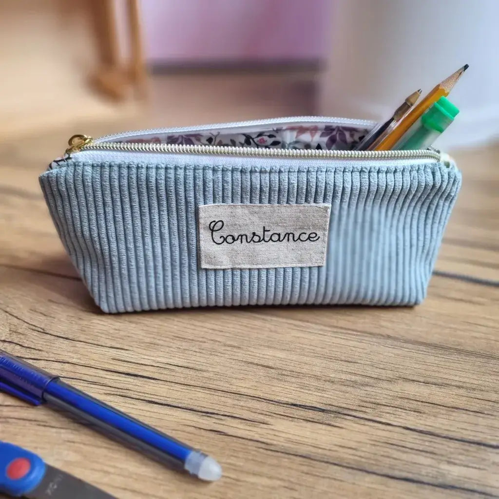 Trousse d’école personnalisée en velours côtelé bleu clair, floquée du prénom "Constance", posée sur un bureau en bois. L’intérieur doublé présente un tissu en coton enduit fleuri visible grâce à la fermeture éclair ouverte. Des crayons, un stylo et un tube de colle dépassent de la trousse. À l’avant, un stylo bleu et des ciseaux pour enfant. Accessoire pratique et élégant pour une rentrée scolaire organisée.