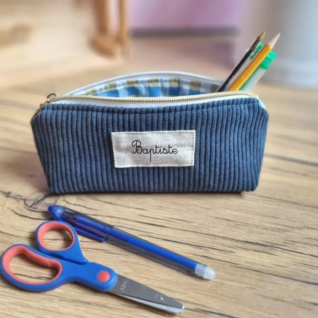 Trousse d’école personnalisée en velours côtelé bleu foncé, floquée du prénom "Baptiste", posée sur un bureau en bois. La trousse est ouverte, laissant entrevoir une doublure en coton enduit à carreaux et des fournitures scolaires : crayons, tube de colle, stylo. À l’avant, une paire de ciseaux pour enfant bleue et rouge et un stylo bille bleu. Accessoire pratique, résistant et élégant pour accompagner les enfants à l’école.