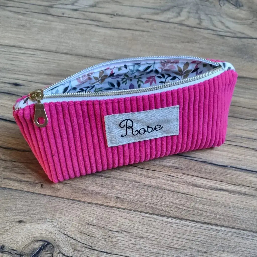 Trousse d’école en velours côtelé framboise, ornée d’une étiquette cousue avec le prénom “Rose” en écriture élégante. Elle est équipée d’une fermeture éclair dorée et doublée d’un tissu intérieur à motif floral multicolore. Posée sur une surface en bois, cette trousse associe style et praticité, idéale pour ranger stylos, crayons et petits accessoires scolaires ou créatifs.