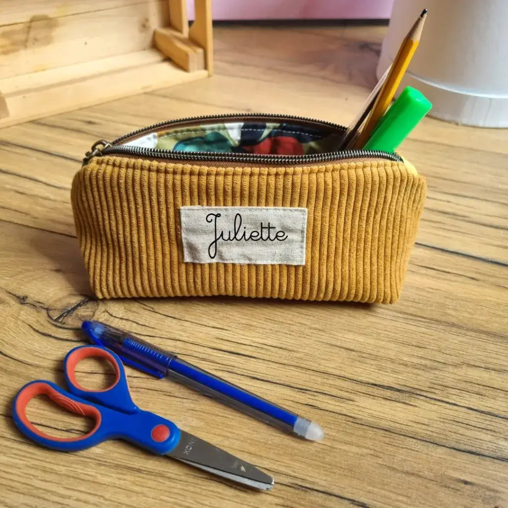 Trousse d’école personnalisée en velours côtelé jaune moutarde, floquée du prénom "Juliette", posée sur un bureau en bois. La trousse est ouverte, laissant apparaître un intérieur en coton enduit fleuri et des fournitures : crayon, feutre vert, tube de colle. Devant, une paire de ciseaux pour enfant bleue et rouge et un stylo bille bleu. Accessoire scolaire tendance et pratique pour accompagner les enfants au quotidien.