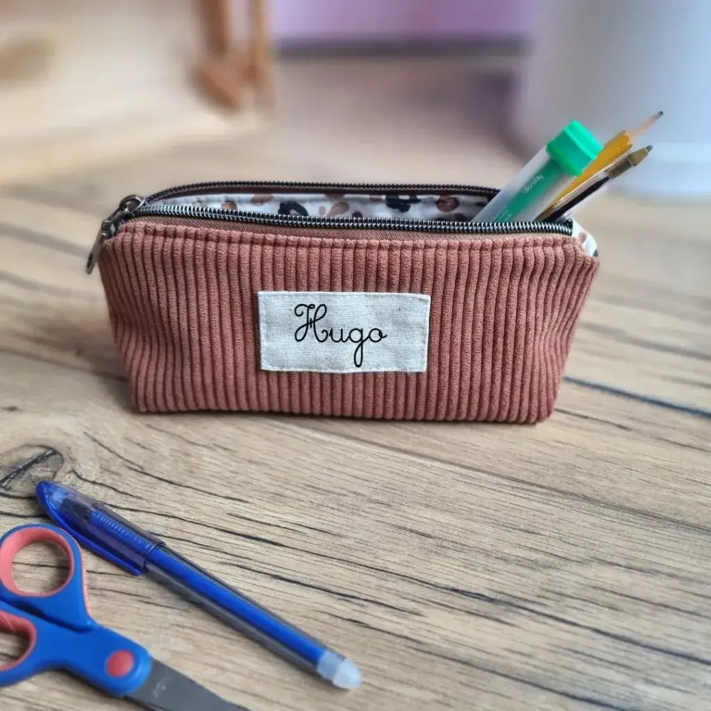 Trousse scolaire en velours côtelé marron personnalisée avec le prénom "Hugo", posée sur un bureau en bois clair. L’intérieur en coton enduit à motifs est partiellement visible grâce à la fermeture zippée ouverte, laissant dépasser un crayon, un feutre vert et un stylo. À l’avant, une paire de ciseaux bleu et rouge ainsi qu’un stylo bille bleu complètent la scène. Idéale pour organiser les fournitures avec style à l’école.
