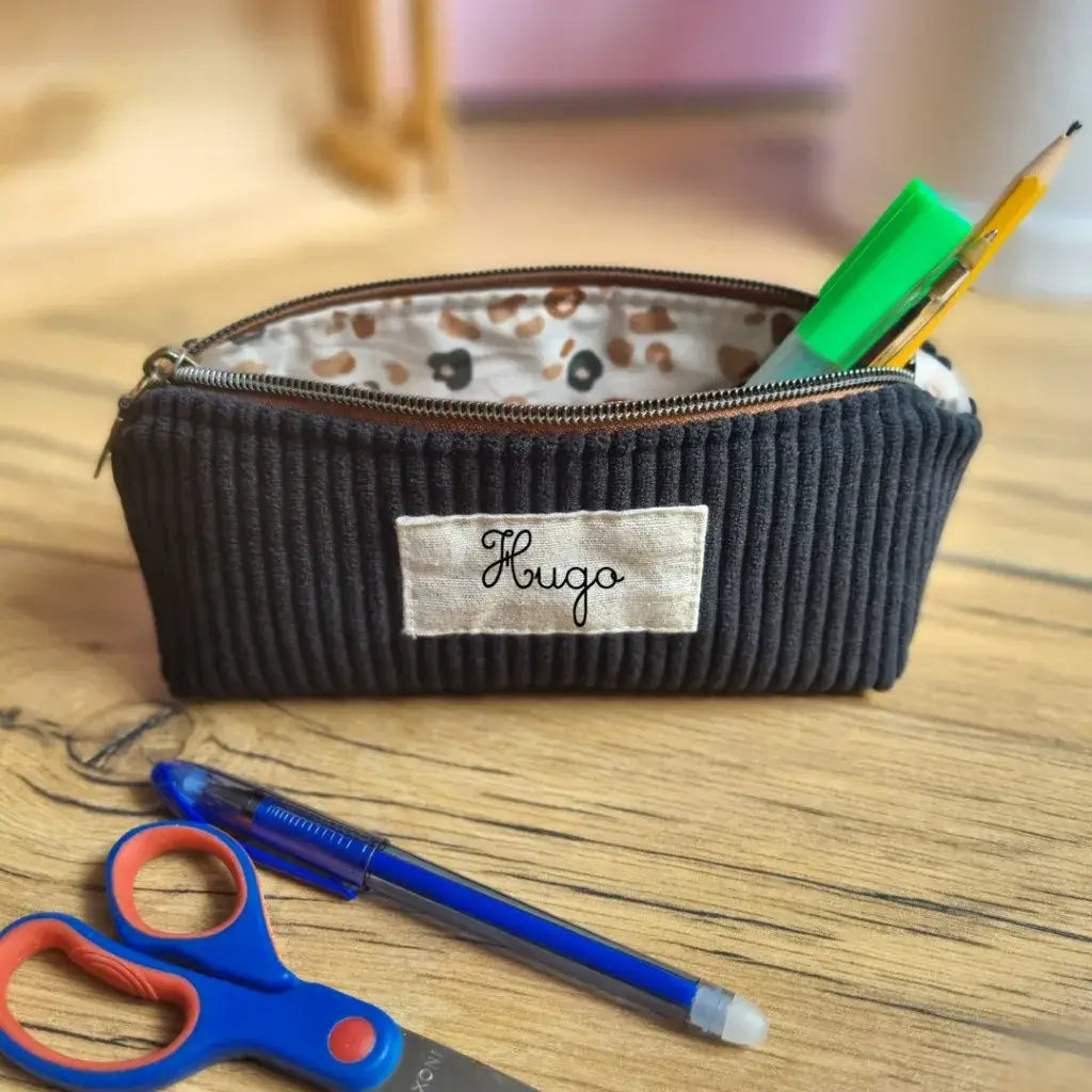 Trousse d’école noire en velours côtelé avec étiquette en tissu floquée du prénom "Hugo", posée sur un bureau en bois. La trousse est ouverte, dévoilant une doublure en coton enduit imprimée d'un motif léopard, et contient un crayon, un feutre vert et un stylo. À l’avant, un stylo bleu et des ciseaux à bouts ronds bleu et rouge. Accessoire élégant et personnalisable pour organiser ses fournitures scolaires avec style.