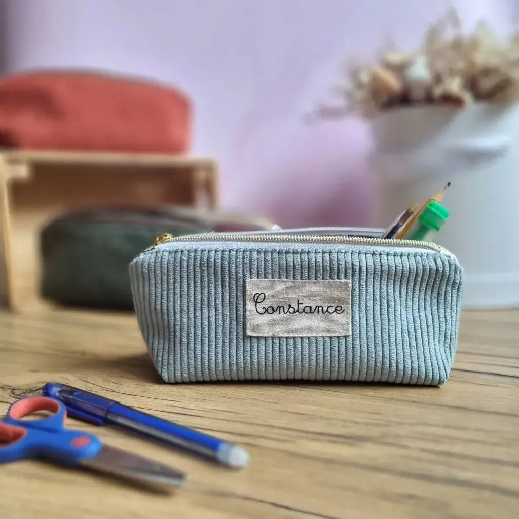 Trousse d’école en velours côtelé bleu clair, personnalisée avec le prénom "Constance" floqué sur une étiquette en tissu. Placée sur un bureau en bois, elle est ouverte et contient un crayon, un feutre et un stylo. En arrière-plan, plusieurs autres trousses de coloris différents sont disposées sur un présentoir en bois. Une paire de ciseaux bleue et un stylo bille sont posés au premier plan. Accessoire scolaire unique, pratique et personnalisable pour la rentrée des classes.
