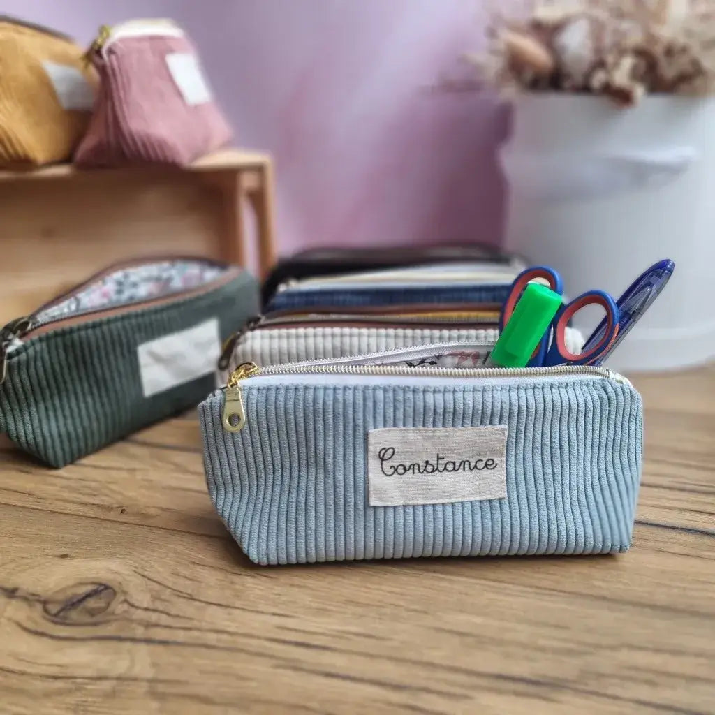 Ensemble de trousses d’école personnalisées en velours côtelé, aux couleurs variées (bleu clair, vert, beige, noir, rose, jaune), posées sur un bureau en bois. Au premier plan, une trousse floquée "Constance", ouverte et garnie de fournitures : feutre vert, ciseaux, stylo. En arrière-plan, plusieurs modèles similaires. Accessoires pratiques et esthétiques pour une rentrée scolaire organisée et stylée.