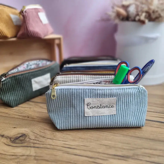 Ensemble de trousses d’école personnalisées en velours côtelé, aux couleurs variées (bleu clair, vert, beige, noir, rose, jaune), posées sur un bureau en bois. Au premier plan, une trousse floquée "Constance", ouverte et garnie de fournitures : feutre vert, ciseaux, stylo. En arrière-plan, plusieurs modèles similaires. Accessoires pratiques et esthétiques pour une rentrée scolaire organisée et stylée.
