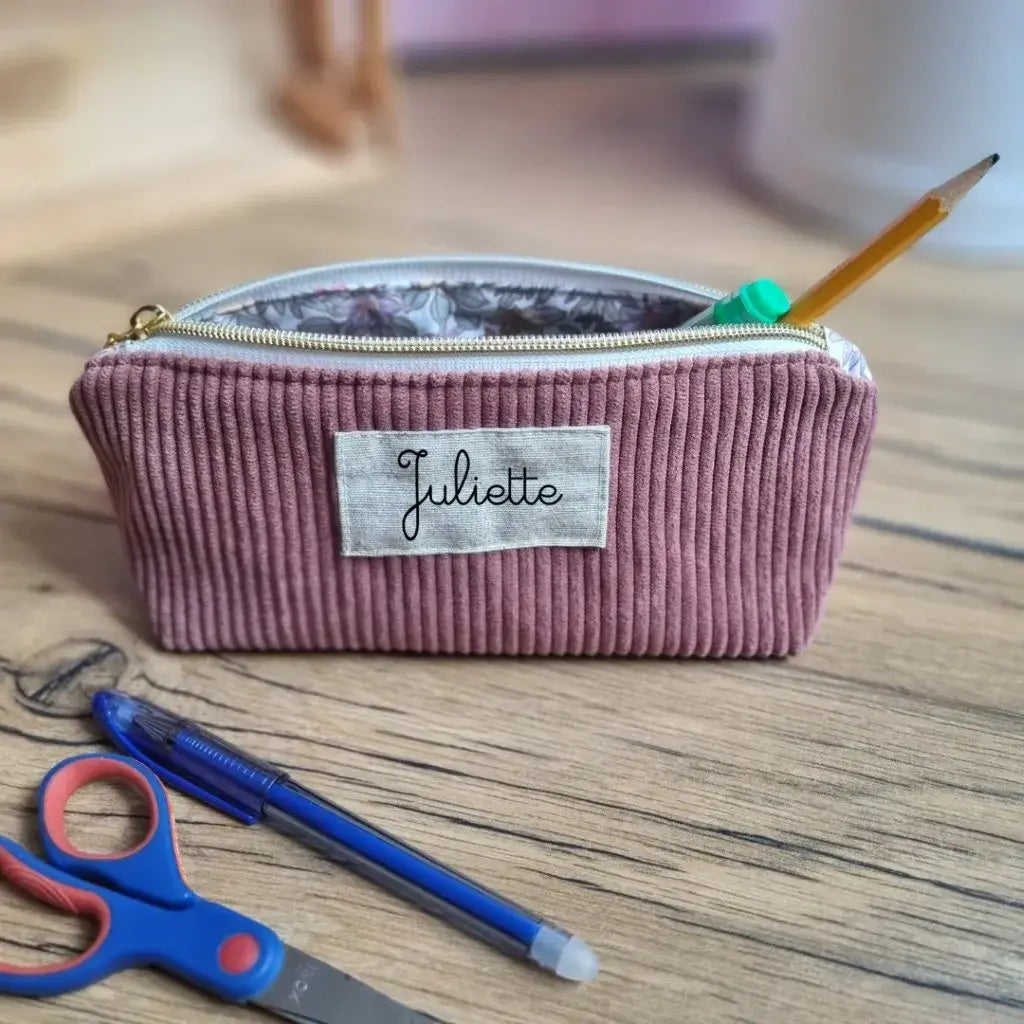 Trousse scolaire personnalisée en velours côtelé rose, floquée du prénom "Juliette", posée sur un bureau en bois clair. Ouverte, elle laisse apparaître une doublure en coton enduit à motif floral et contient un crayon, un feutre vert et un stylo. Devant la trousse, une paire de ciseaux bleu et rouge ainsi qu’un stylo bille bleu. Accessoire original et pratique pour bien préparer la rentrée des classes.