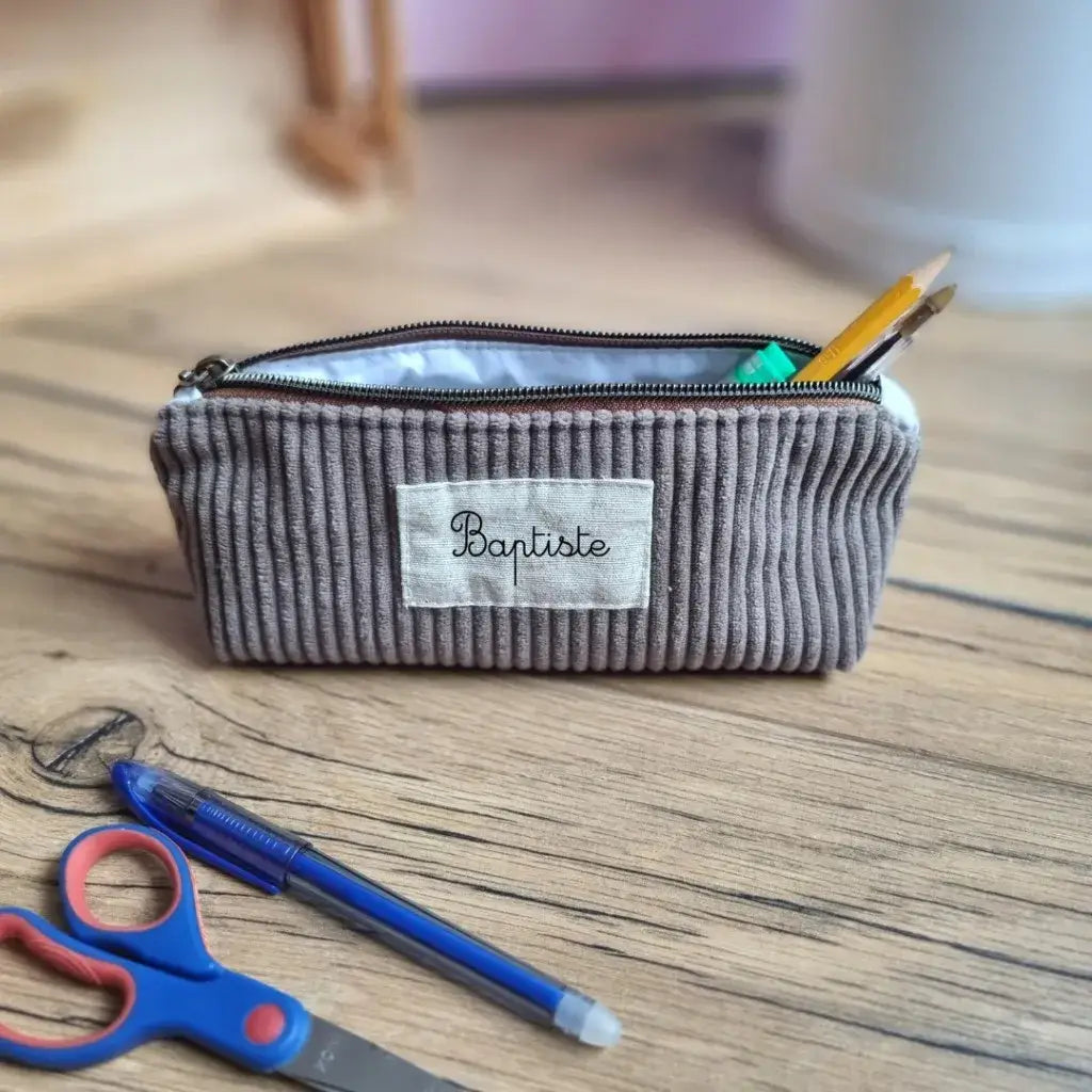 Trousse d’école en velours côtelé taupe personnalisée avec le prénom "Baptiste", posée sur un bureau en bois. Ouverte, elle dévoile un intérieur en coton enduit uni blanc, contenant un crayon, un feutre vert et un stylo. À l’avant, une paire de ciseaux bleu et rouge ainsi qu’un stylo bille bleu sont visibles. Accessoire scolaire chic et fonctionnel, parfait pour ranger ses fournitures avec soin.