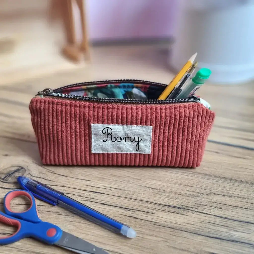 Trousse d’école en velours côtelé terracotta personnalisée avec le prénom "Romy", posée sur un bureau en bois clair. L’intérieur en coton enduit à motif coloré est visible grâce à la fermeture zippée ouverte, laissant dépasser un crayon, un feutre vert et un stylo. Devant la trousse, une paire de ciseaux pour enfant bleue et rouge ainsi qu’un stylo bille bleu. Accessoire tendance et utile pour une organisation stylée des fournitures scolaires.