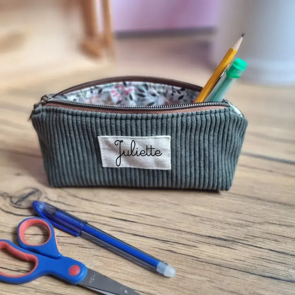 Trousse scolaire en velours côtelé vert avec étiquette personnalisée "Juliette", posée sur un sol en bois clair. Son ouverture dévoile un tissu intérieur en coton enduit à motifs floraux délicats, accompagnée de fournitures scolaires visibles : crayon, feutre vert et stylo. Un stylo bille bleu et des ciseaux à bouts ronds reposent juste devant. Une trousse raffinée, idéale pour une rentrée organisée et stylée.
