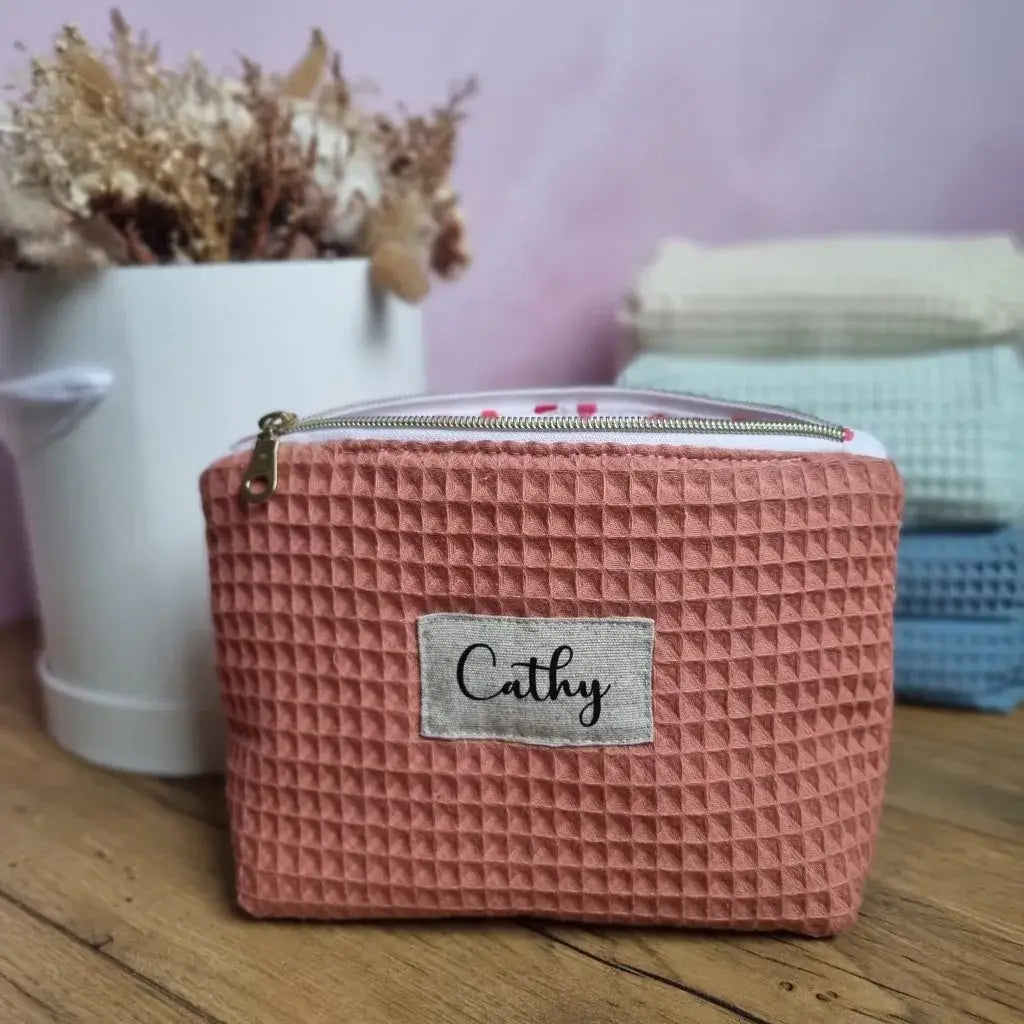 Au premier plan, une trousse zippée en tissu nid d'abeille de coloris marsala personnalisée avec le prénom "Cathy" floquée sur une étiquette en toile. A l'arrière-plan, trois autres trousses similaires de coloris naturel, menthe et bleu denim sont empilées les unes sur les autres à côté d'un bouquet de fleurs séchées.