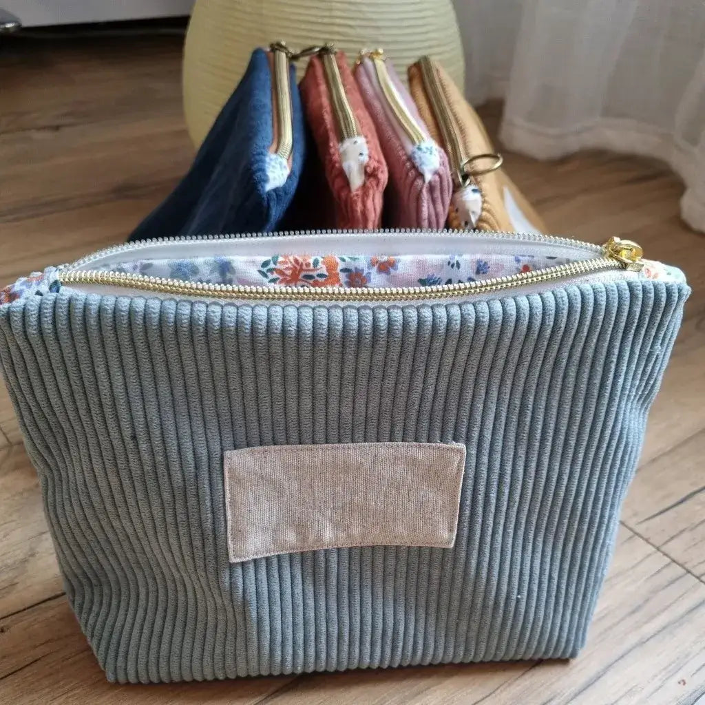 Trousse zippée en velours côtelé bleu clair avec une doublure intérieure en coton imprimé à motif fleuri. Cette trousse dispose d'une étiquette en tissu cousue en façade pouvant accueillir un prénom ou une inscription personnalisée qui sera floquée dessus. A l'arrière plan, on distingue quatre autres trousses zippées en velours côtelé du même type et de coloris divers disposées les unes contre les autres devant une lampe.