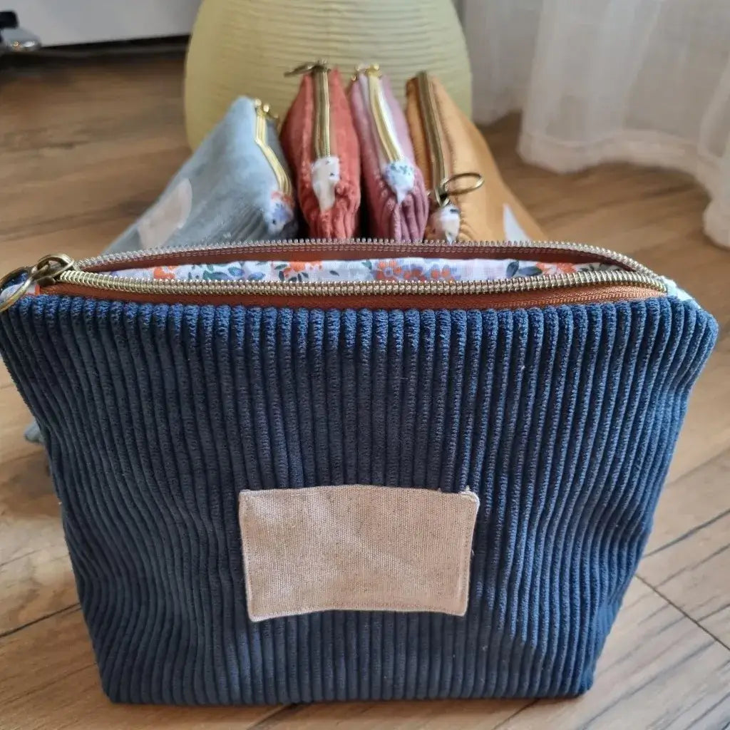 Trousse zippée en velours côtelé bleu foncé avec une doublure intérieure en coton imprimé à motif fleuri. Cette trousse dispose d'une étiquette en tissu cousue en façade pouvant accueillir un prénom ou une inscription personnalisée qui sera floquée dessus. A l'arrière plan, on distingue quatre autres trousses zippées en velours côtelé du même type et de coloris divers disposées les unes contre les autres devant une lampe.