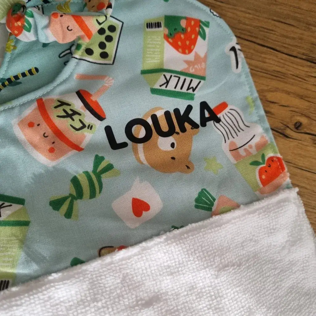 Zoom sur la partie en tissu coton imprimé à motif et sur la personnalisation au prénom d'un bavoir cantine élastique pour maternelle. Le bavoir présenté est au motif Kawai et est personnalisé avec le prénom "Louka" floqué sur la partie en coton avec une écriture majuscule noire.