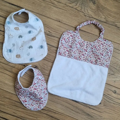 Ensemble de trois bavoirs bébé faits main, présentés sur un sol en bois. Le lot comprend un bavoir classique à motifs d’animaux (lion, girafe, éléphant), un bavoir bandana et un grand bavoir à enfilage facile, tous deux assortis d’un tissu fleuri rouge et rose. Chaque modèle est doublé en tissu éponge blanc ultra-absorbant, offrant confort et protection lors des repas. Un lot pratique, évolutif et esthétique pour accompagner bébé au quotidien.