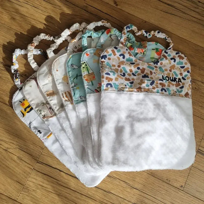 Sept bavoirs cantines élastiques pour maternelle en éponge de bambou blanc et tissu imprimé de motifs divers sont disposés en éventail au sol sur un parquet en chêne. Le premier bavoir est personnalisé avec le prénom "Louka" floqué dessus en écriture noire.