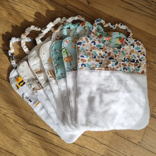 Sept bavoirs cantines élastiques pour maternelle en éponge de bambou blanc et tissu imprimé de motifs divers sont disposés en éventail au sol sur un parquet en chêne. Le premier bavoir est personnalisé avec le prénom "Louka" floqué dessus en écriture noire.