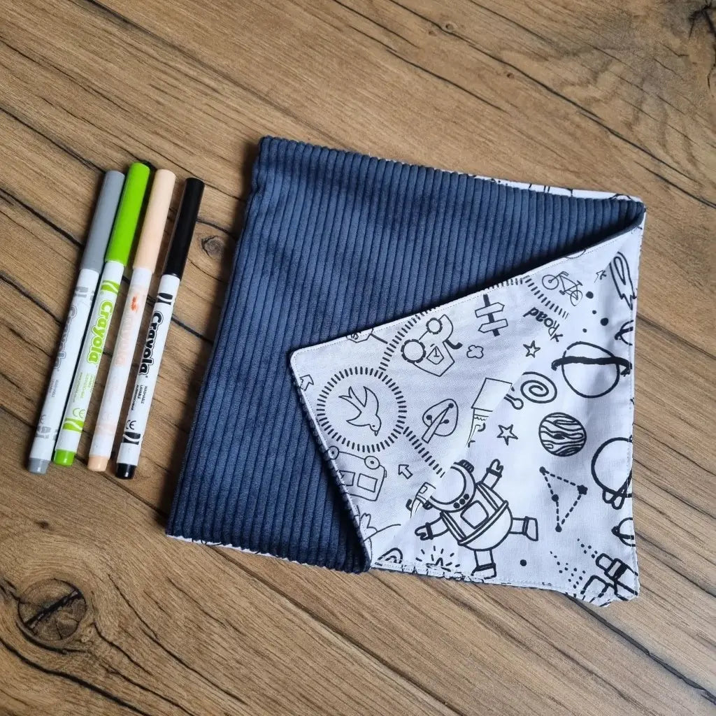 Cahier de coloriage en tissu avec une couverture en velours bleu. Le coin inférieur droit est replié sur lui-même montrant une partie des motifs à colorier. Quatre feutres de couleurs sont alignés à la gauche de ce cahier en tissu.