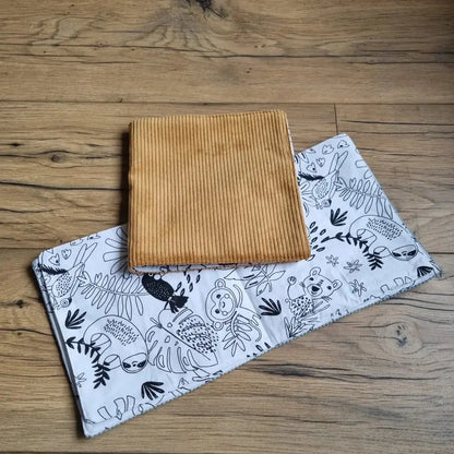 Deux cahiers de coloriage en tissu posés au sol sur un parquet en chêne. Celui du dessus est fermé. Sa couverture est en velours côtelé de couleur jaune. Celui du dessous est ouvert sur une double page de motifs savane à colorier.