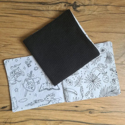 Deux cahiers de coloriage en tissu posés au sol sur un parquet en chêne. Celui du dessus est fermé. Sa couverture est en velours côtelé de couleur noire. Celui du dessous est ouvert sur une double page de motifs divers à colorier.