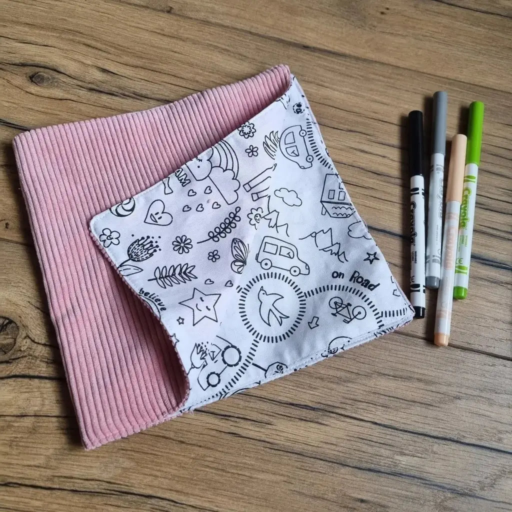 Cahier de coloriage en tissu avec une couverture en velours côtelé rose. Le coin inférieur droit est replié pour montrer quelques exemples de motifs à colorier à l'intérieur de ce cahier. Quatre feutres de couleurs sont alignés à droite de ce cahier en tissu.