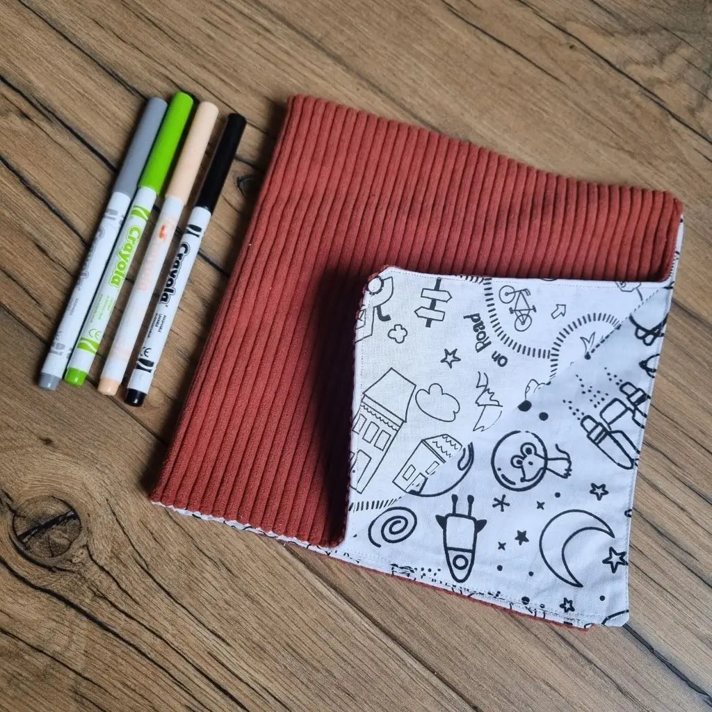 Cahier de coloriage en tissu lavable et réutilisable avec une couverture de couleur terracotta. Le coin inférieur droit est replié sur lui-même pour montrer quelques motifs à colorier. Quatre feutres de couleurs sont disposés à la gauche de ce cahier en tissu.