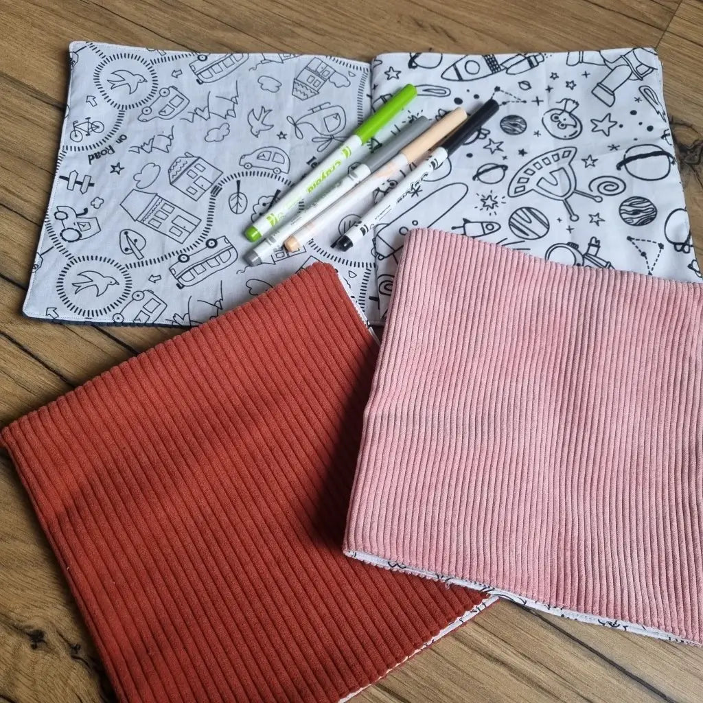 Trois cahiers de coloriage en tissu, celui du haut ouvert sur une double page à colorier avec quatre feutres de couleurs posés dessus, les deux en dessous couverture fermée, celui de gauche avec une couverture terracotta, celui de droite avec une couverture rose