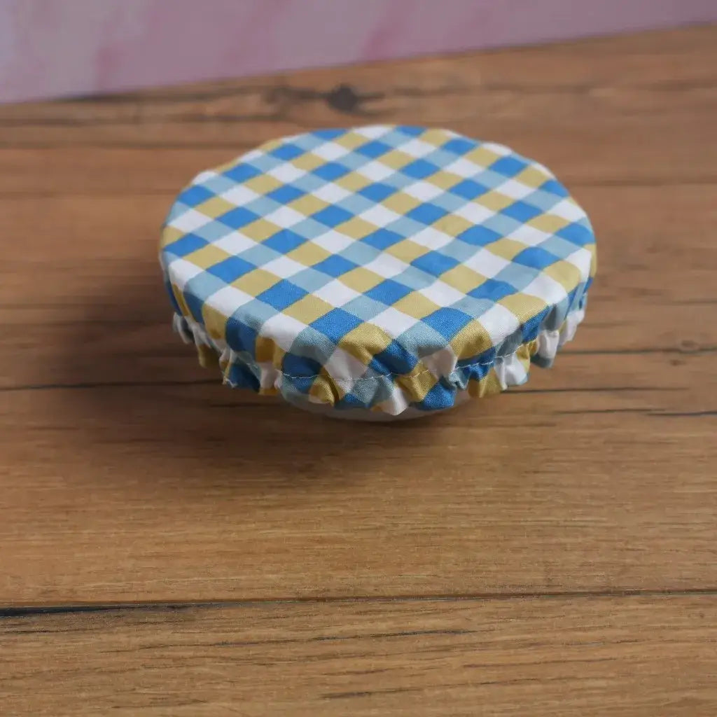 Petite charlotte couvre-plat ronde en coton enduit à motif carreaux bleus et jaunes, format taille S, réutilisable et lavable, dotée d’un élastique froncé pour s’ajuster sur un bol ou ramequin. Alternative zéro déchet aux films alimentaires pour conserver les restes ou protéger un dessert maison.