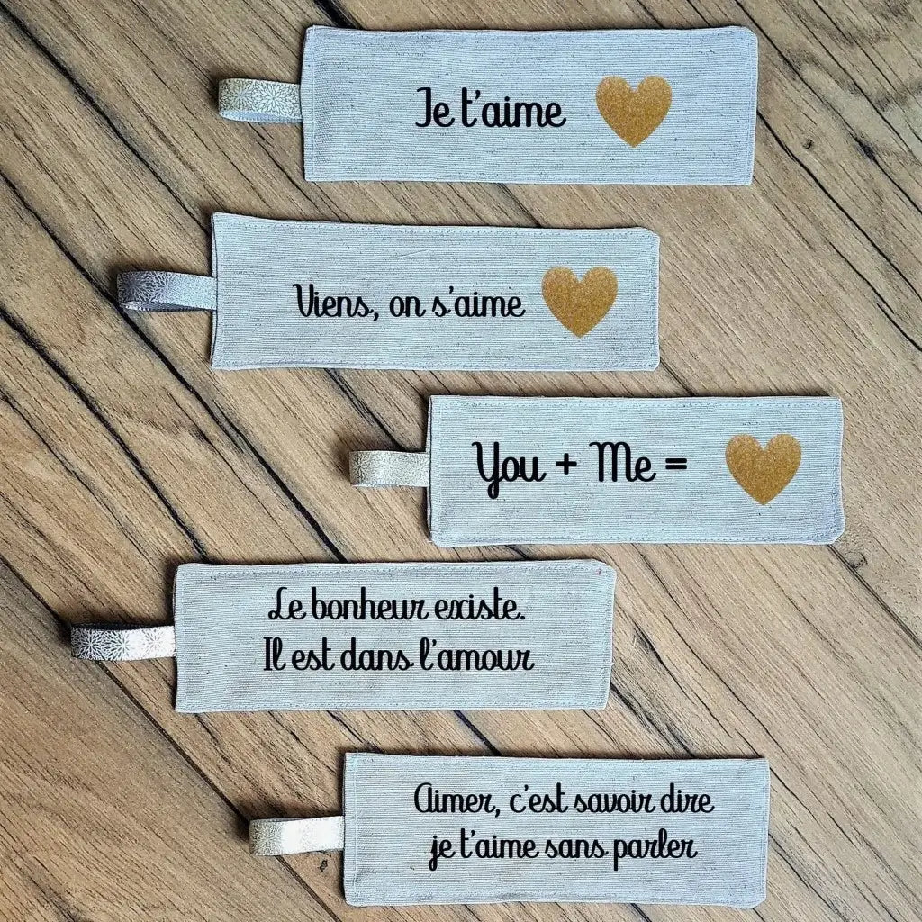 Cinq marque-pages en tissu posés au sol les uns au-dessus des autres sur un parquet en chêne. Tous ces marque-pages en tissu sont personnalisés avec une inscription floquée dessus. "Je t'aime", "Viens, on s'aime", "You + Me", "Le bonheur existe. Il est dans l'amour", "Aimer, c'est savoir dire je t'aime sans parler".