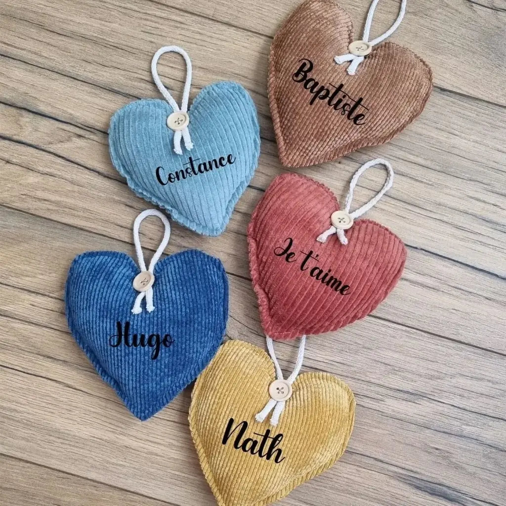 Cinq cœurs en tissu velours côtelé posés sur un parquet en chêne. De haut en bas : couleur marron avec le prénom "Baptiste", couleur bleu clair avec le prénom "Constance", couleur terracotta avec l'inscription "Je t'aime", couleur bleu foncé avec le prénom "Hugo", couleur jaune avec le prénom "Nath".