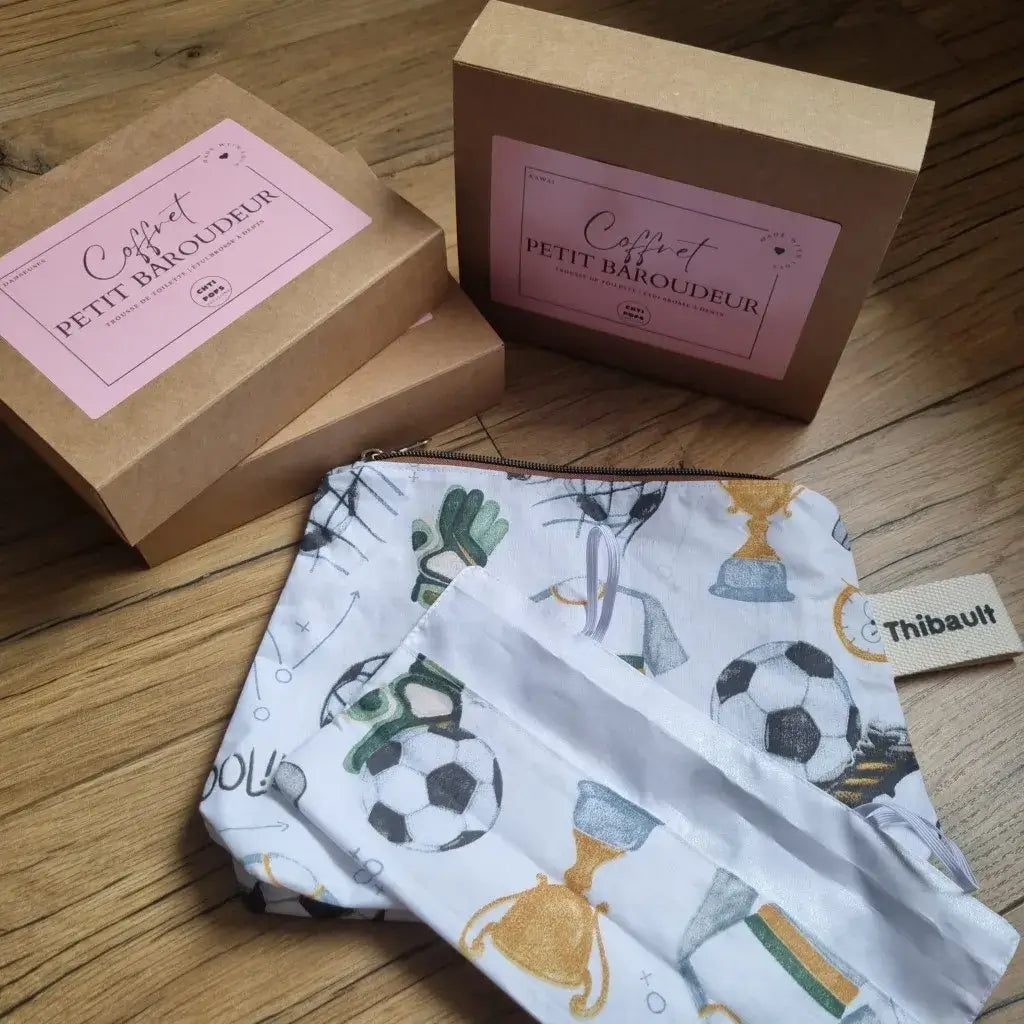 Coffret "petit baroudeur" par Chtipops Créations. Ce coffret est composé d'une trousse de toilette imperméable à fermeture zippée ainsi que d'un étui à brosse à dents en tissu se fermant grâce à deux petits élastiques. Le motif présenté ici est le motif "foot". A l'arrière plan, les boîtes en carton contenant ces kits "petit baroudeur".