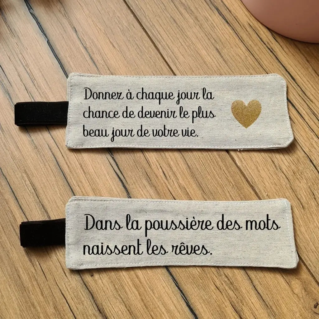 Deux marque-pages en tissu posés au sol sur un parquet en chêne. Ces deux marque-pages en tissu sont personnalisés avec une citation floquée en noir dessus. Pour celui du haut : "Donnez à chaque jour la chance de devenir le plus beau jour de votre vie". Pour celui du bas : "Dans la poussière des mots naissent les rêves".