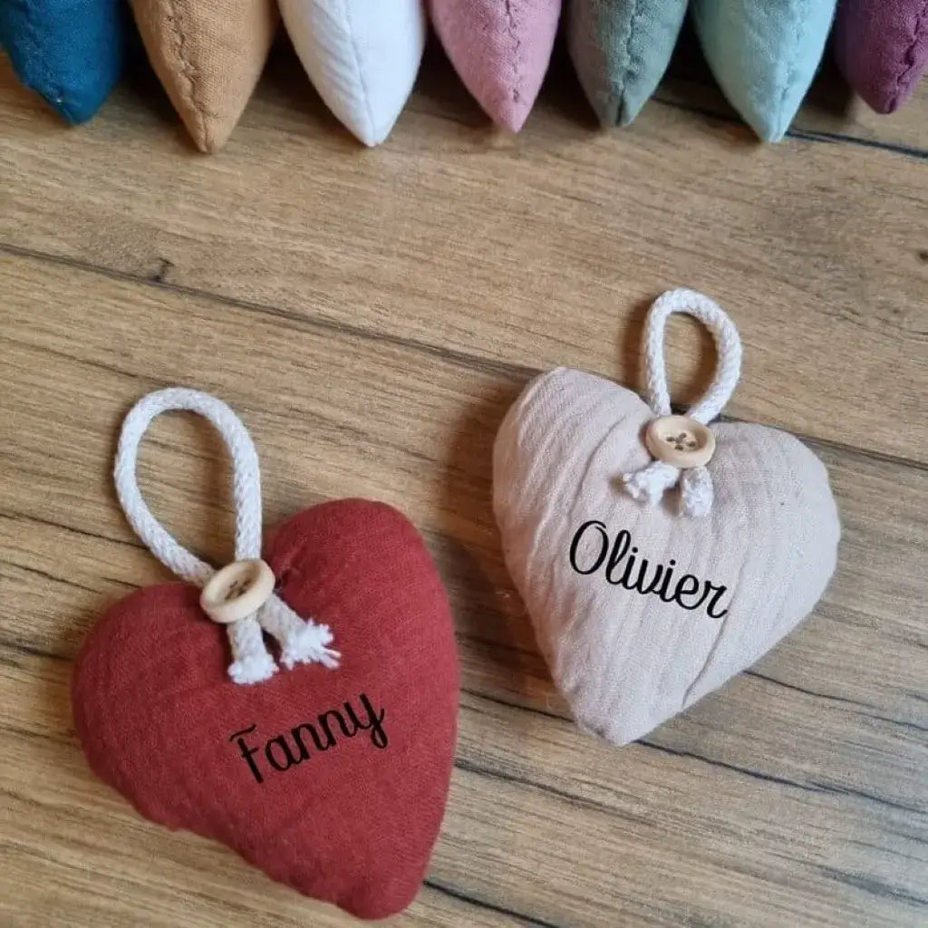 Deux petits cœurs en tissu double gaze de coton posés sur un parquet en chêne. Celui de gauche est de coloris terracotta et comporte l'inscription du prénom "Fanny". Celui de droite est de couleur sable et comporte l'inscription du prénom "Olivier". Sur le dessus de l'image sont présentés l'ensemble des 7 autres coloris disponibles.