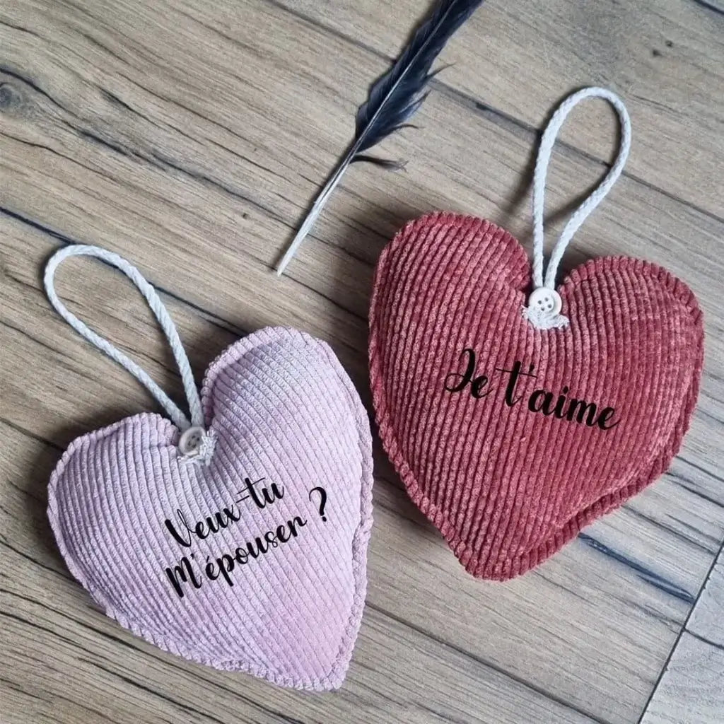 Deux cœurs en tissu velours côtelé posé sur un parquet en chêne. Celui de gauche est de couleur rose et comporte l'inscription "Veux-tu m'épouser ?". Celui de droite est de couleur terracotta et comporte l'inscription "Je t'aime".