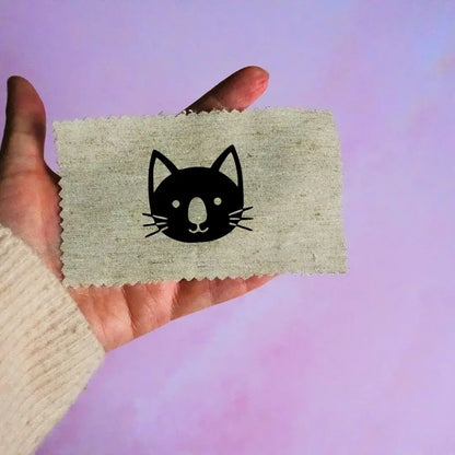 Main tenant une étiquette en tissu beige avec un motif de tête de chat noir minimaliste, sur fond lavande.