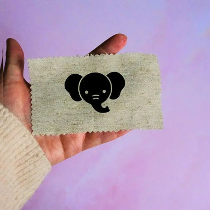 Main tenant une étiquette en tissu beige avec un motif de tête d'éléphant noir minimaliste, sur fond lavande.