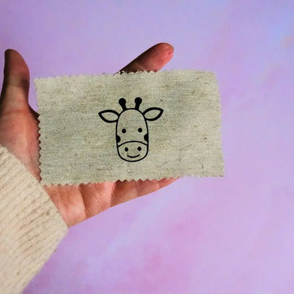 Main tenant une étiquette en tissu beige avec un motif de tête de girafe noir minimaliste, sur fond lavande.