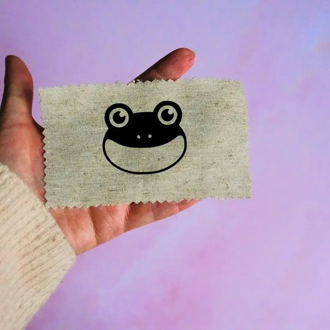 Main tenant une étiquette en tissu beige avec un motif de tête de grenouille noir minimaliste, sur fond lavande.