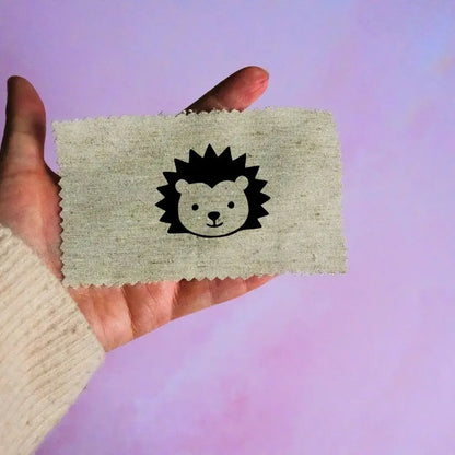 Main tenant une étiquette en tissu beige avec un motif de tête de hérisson noir minimaliste, sur fond lavande.