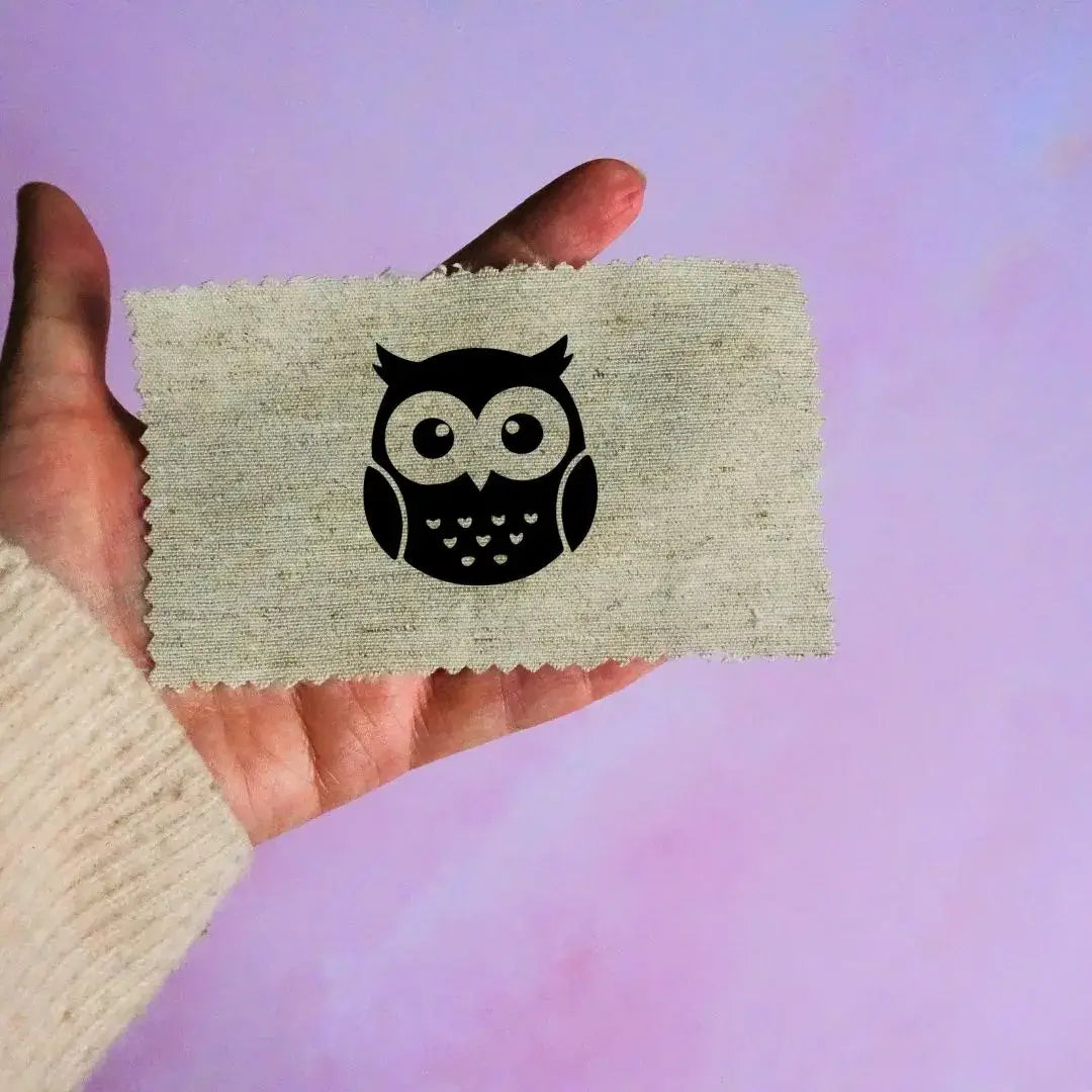 Main tenant une étiquette en tissu beige avec un motif de tête de hibou noir minimaliste, sur fond lavande.