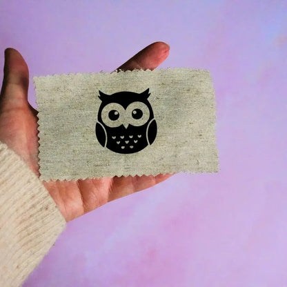 Main tenant une étiquette en tissu beige avec un motif de tête de hibou noir minimaliste, sur fond lavande.