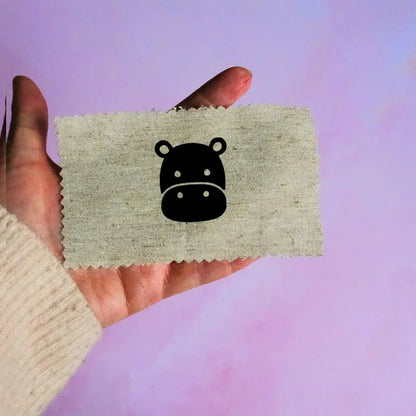 Main tenant une étiquette en tissu beige avec un motif de tête d'hippopotame noir minimaliste, sur fond lavande.