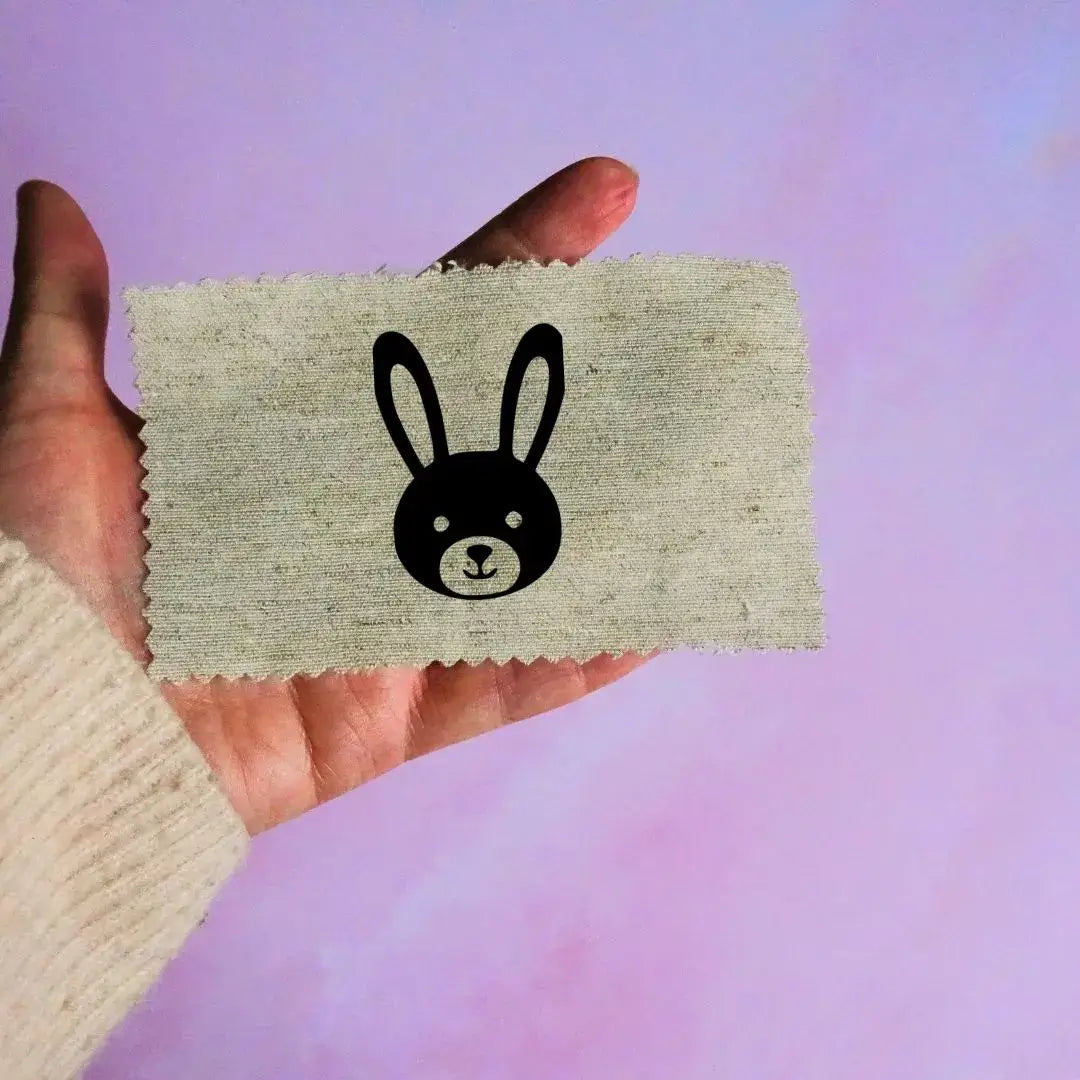 Main tenant une étiquette en tissu beige avec un motif de tête de lapin noir minimaliste, sur fond lavande.
