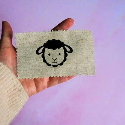 Main tenant une étiquette en tissu beige avec un motif de tête de mouton noir minimaliste, sur fond lavande.