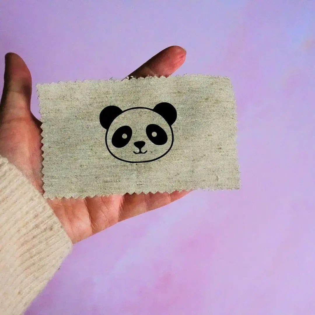 Main tenant une étiquette en tissu beige avec un motif de tête de panda noir minimaliste, sur fond lavande.
