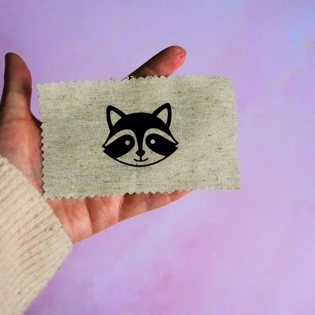 Main tenant une étiquette en tissu beige avec un motif de tête de raton laveur noir minimaliste, sur fond lavande.