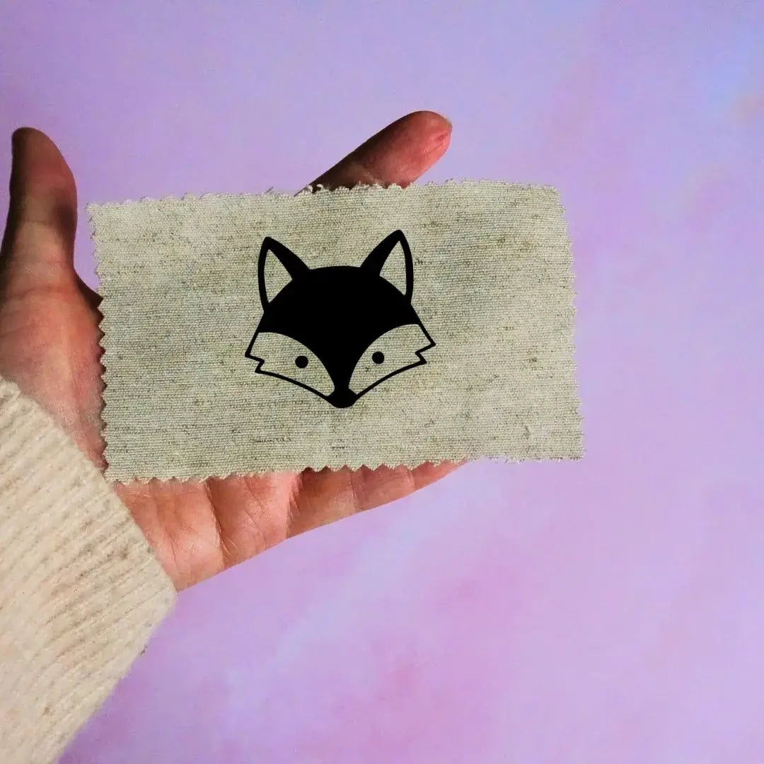 Main tenant une étiquette en tissu beige avec un motif de tête de renard noir minimaliste, sur fond lavande.