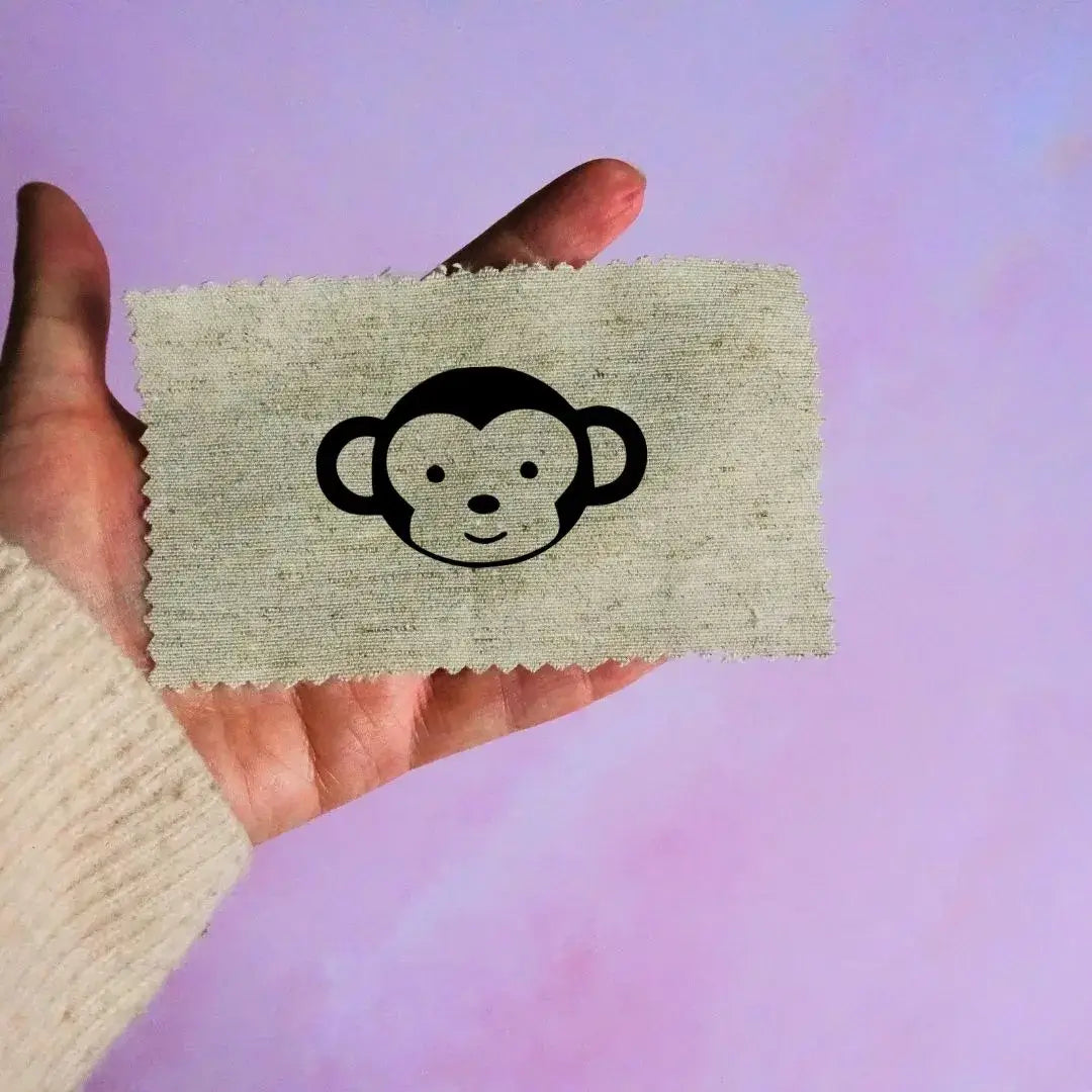 Main tenant une étiquette en tissu beige avec un motif de tête de singe noir minimaliste, sur fond lavande.